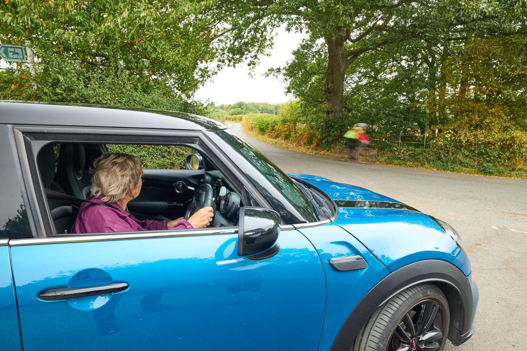 A mature driver in a blue Mini on a wooded lane